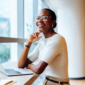 istockphoto-2219635385-612×612 A cheerful young black woman wearing glasses is sitting by a window with a laptop, confidently exuding professionalism and positivity. She appears approachable and engaged in her workplace setting.